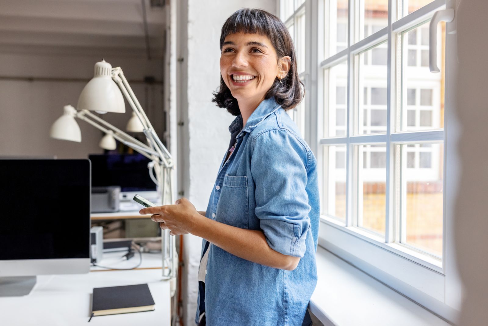 Young businesswoman standing by the window with mobile phone at startup office. Female professional taking break from work at office.